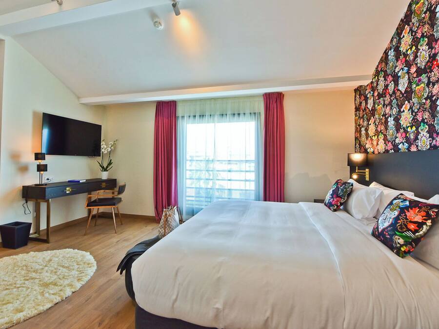 Quarto de hotel com uma cama grande, uma escrivaninha com televisão, uma janela com cortinas e uma tapeçaria colorida na parede.