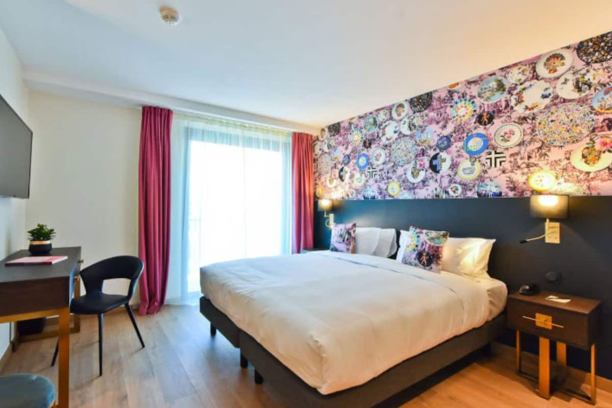 Una habitación de hotel con cama, escritorio, silla, televisor y ventana con cortinas.