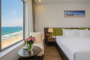 A hotel room with a bed, a chair, a small table with a vase of flowers, a nightstand with a lamp, and a painting on the wall.