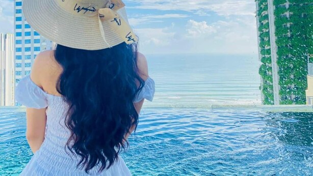 A person in a white dress and hat overlooking a body of water with tall buildings in the background. - Santa Luxury Hotel (Da Nang)