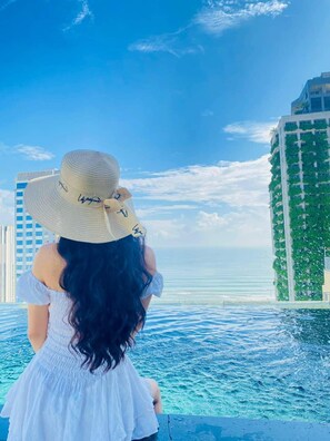 A person in a white dress and hat overlooking a body of water with tall buildings in the background. - Santa Luxury Hotel (Da Nang)