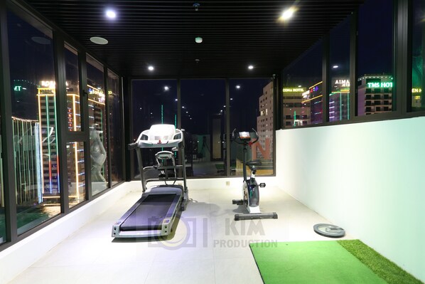 Un gimnasio con equipo de ejercicio, ventanas amplias y vista a edificios de la ciudad de noche.