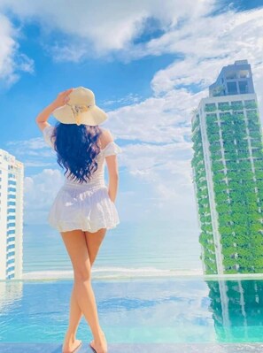 A woman in a white dress and hat standing by a pool with a view of a tall building and the ocean. - Santa Luxury Hotel (Da Nang)
