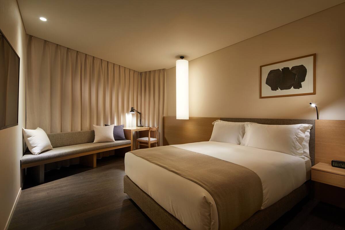 A hotel room with a large bed, a bench with pillows, a desk with a lamp, and a wall-mounted artwork.