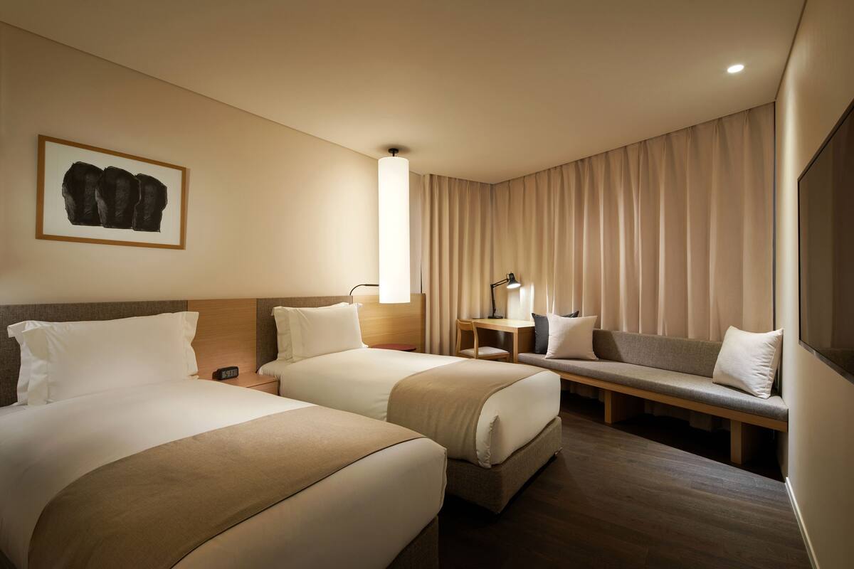 A hotel room with two beds, a desk, a bench, and a large window with curtains.
