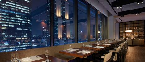 Un restaurante con mesas preparadas para comer, amplios ventanales que ofrecen vistas de la ciudad y una zona de bar con estantes llenos de botellas.