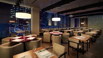 A modern restaurant with tables set for dining, large windows offering city views, and contemporary lighting fixtures.