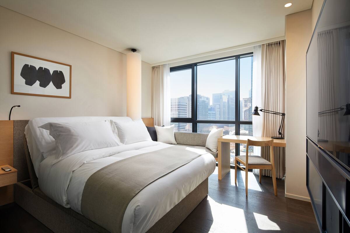 A hotel room with a large bed, a desk, and a chair. There is a window with a city view.