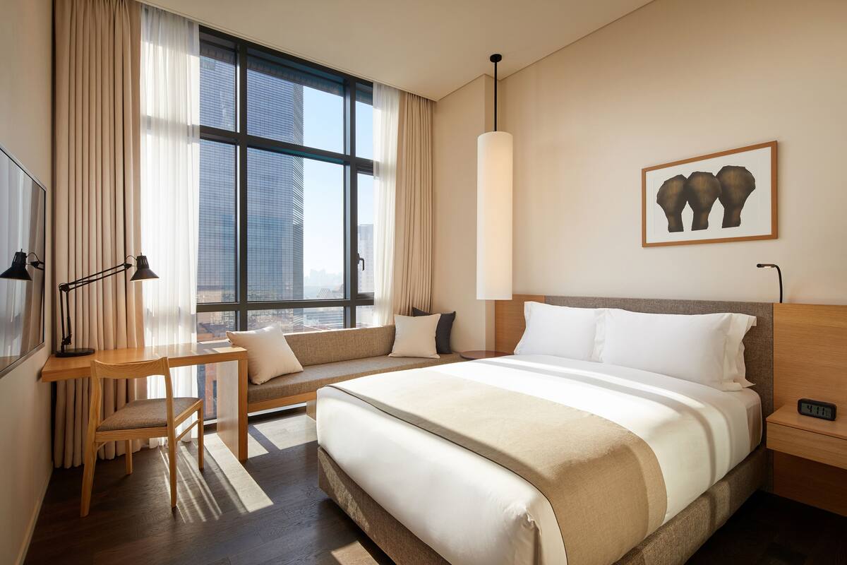A hotel room with a large bed, a desk with a chair, a window with curtains, a wall-mounted artwork, and a nightstand with a lamp.