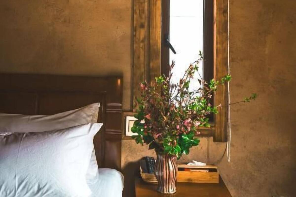 A bedroom with a wooden headboard, a bedside table with a vase of flowers, a window with a wooden frame, and a hanging light fixture.