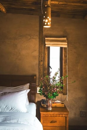 A bedroom with a wooden headboard, a bedside table with a vase of flowers, a window with a wooden frame, and a hanging light fixture. - Guyue Caotang (Huzhou)