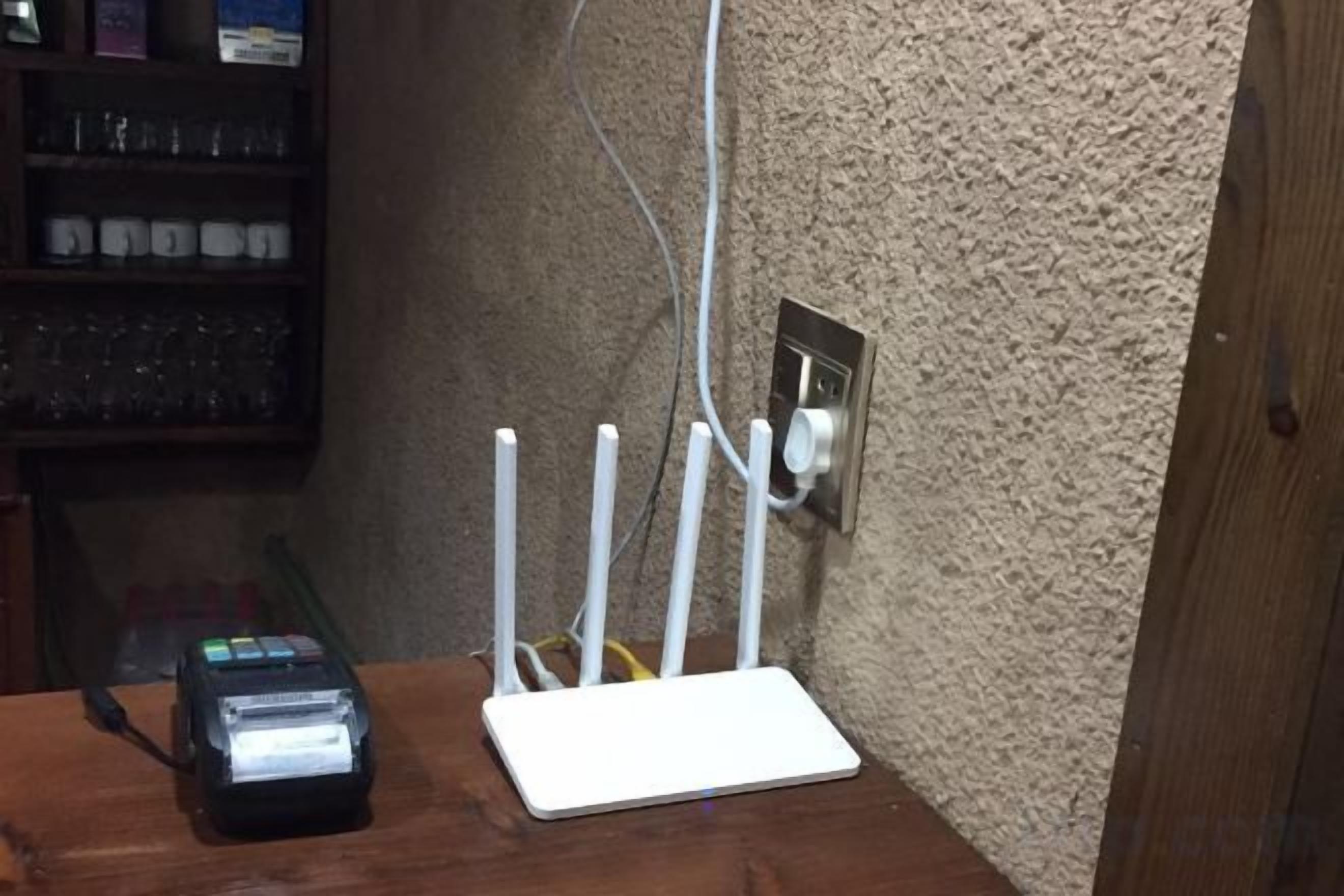 Un router blanco sobre una superficie de madera, con un lector de códigos de barras a su lado, una pared con textura y un enchufe.