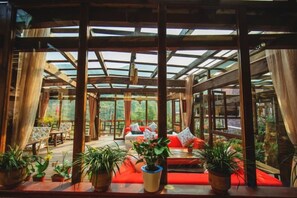 A covered outdoor seating area with red cushions, potted plants, and a glass ceiling structure. - Guyue Caotang (Huzhou)
