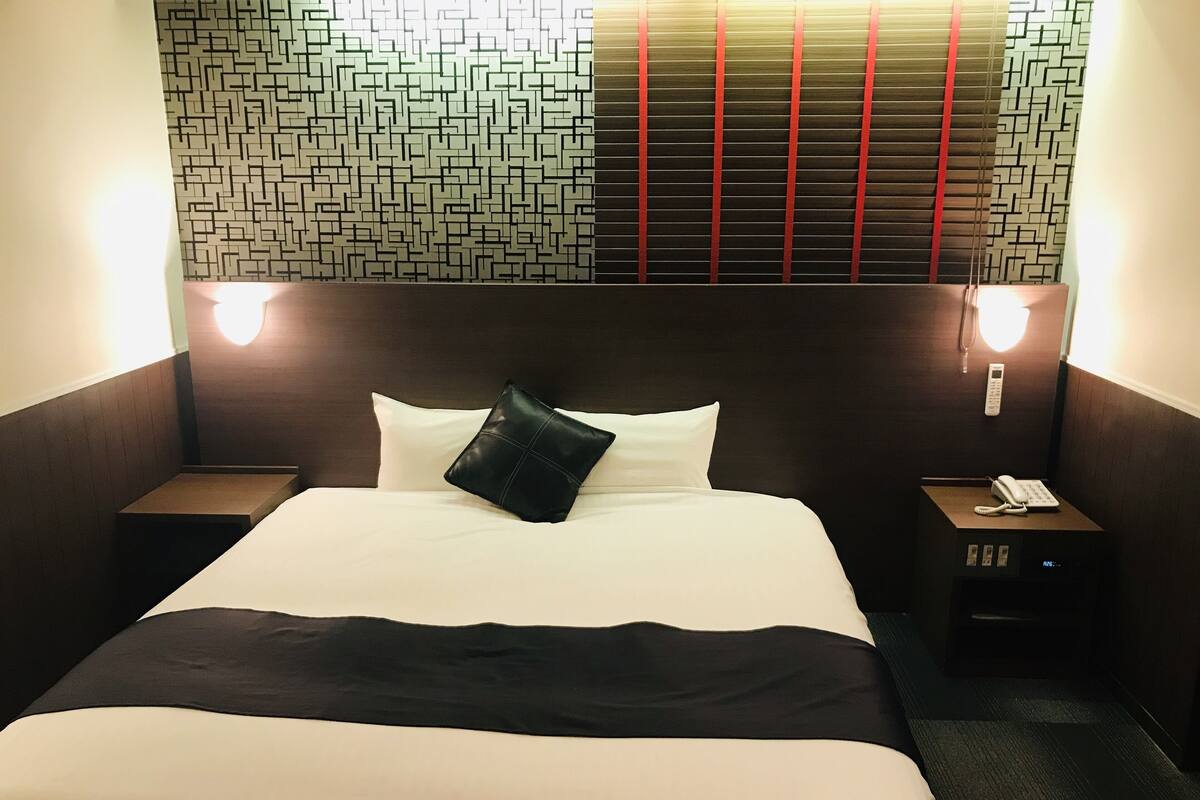 A hotel room with a neatly made bed, a bedside table with a phone, and a wall with a geometric pattern.