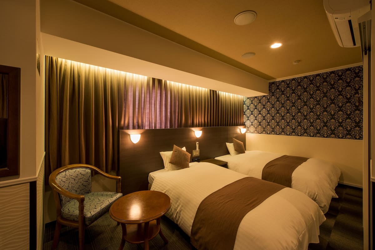 A hotel room with two beds, a chair, a small table, and a headboard with wall-mounted lights.