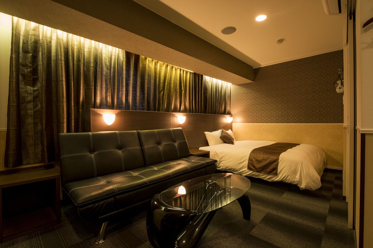 A hotel room with a black leather sofa, a glass coffee table, a bed with white bedding, and a wall with a curtain.