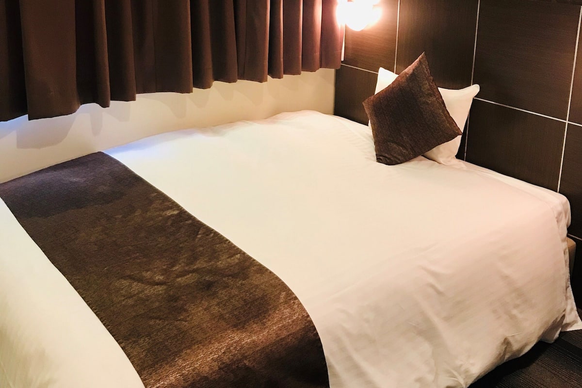 A hotel room with a large bed, dark curtains, a headboard with a patterned design, and a bedside lamp.