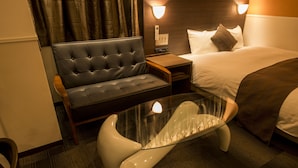 A modern hotel room with a tufted leather sofa, a glass-top coffee table, a bedside table with a lamp, and a neatly made bed. - Sejour Inn Nagarekawa (Hiroshima)