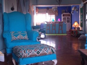 A living room with a large blue armchair and ottoman, a wooden dining table with chairs, a kitchen area in the background, and a small table with a lamp.