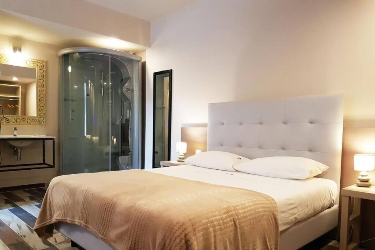 Habitación de hotel con una cama grande, ducha con mampara de vidrio, tocador con espejo y mesita de noche con lámpara.