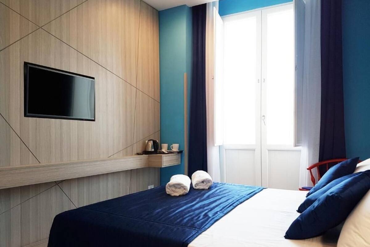Una habitación de hotel con cama, televisor de pantalla plana montado en la pared, ventana con cortinas y una pequeña estantería con objetos.