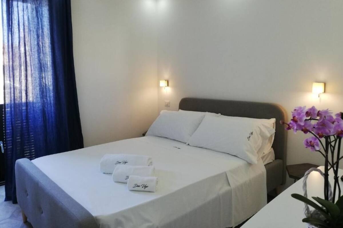 Una cama bien hecha con sábanas blancas y dos toallas, una mesita de noche con un florero, y una ventana con cortinas azules.