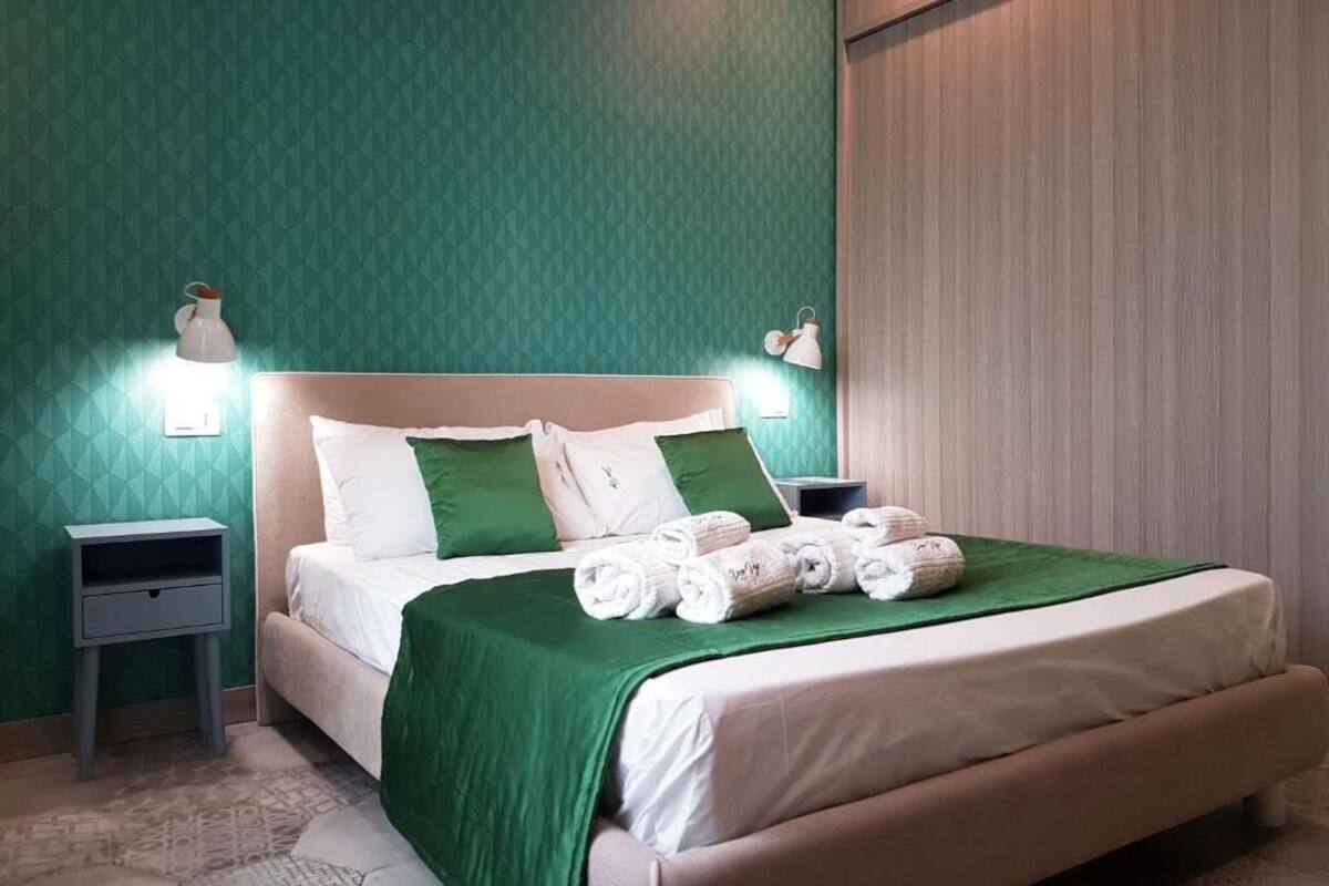 Una cama bien hecha con ropa de cama verde y almohadas blancas, una mesita de noche con una lámpara, y una pared con un diseño geométrico.