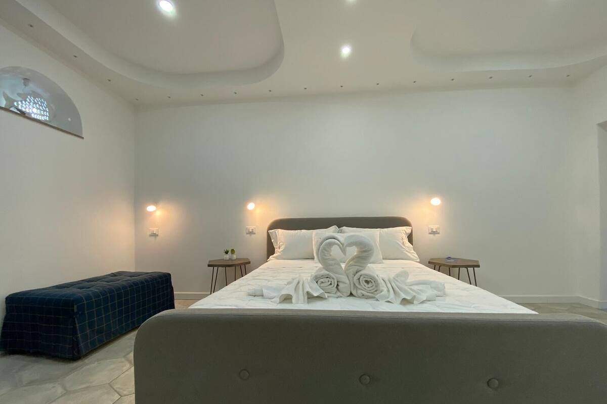 Una habitación de hotel moderna con una cama grande, dos mesitas de noche, un banco y un techo curvo con iluminación empotrada.