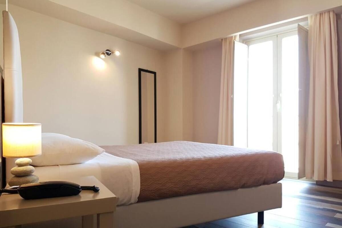 Una habitación de hotel con cama, lámpara de noche y vistas al exterior por una ventana.