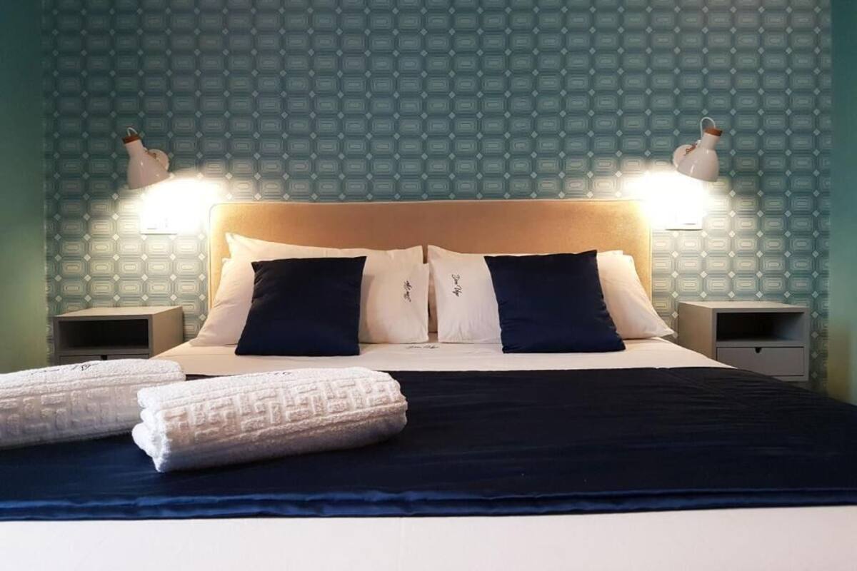 Una cama bien hecha con un edredón azul oscuro, almohadas blancas y un cabecero de madera. Hay dos lámparas de noche a cada lado de la cama.