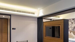 Une chambre d’hôtel moderne avec une table basse ronde, deux fauteuils, un téléviseur posé sur un meuble en bois et un lit doté d’une tête de lit à motifs.