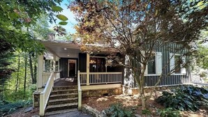 A two-story house with a stone staircase leading to a porch, surrounded by trees and a garden.
