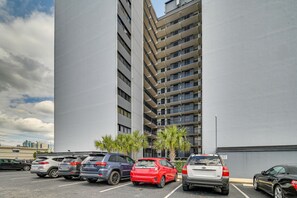 A parking lot with several cars, a tall building with multiple balconies, and some palm trees. - Walk to Family Kingdom, Ocean: Myrtle Beach Condo! (Myrtle Beach)