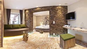Ein moderner Wellnessraum mit Steinwand, einer Massageliege, einer grünen gepolsterten Bank, einem Glastisch und einer Holzbank.