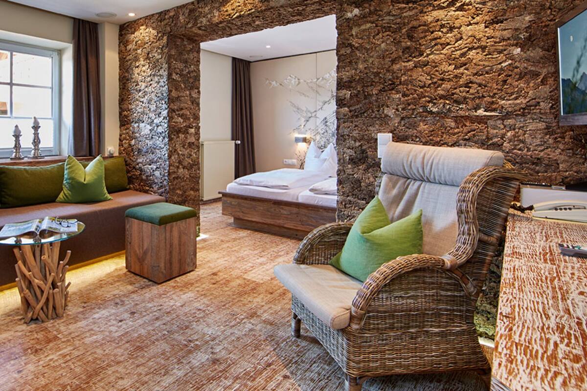 A cozy room with a stone wall, a wooden door, a sofa with green cushions, a wicker chair, and a small table with a magazine.