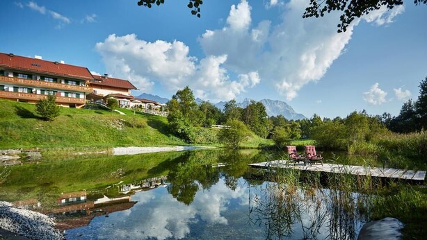 Blick auf einen See mit einem Gebäude, einem Steg und Stühlen am Wasser.