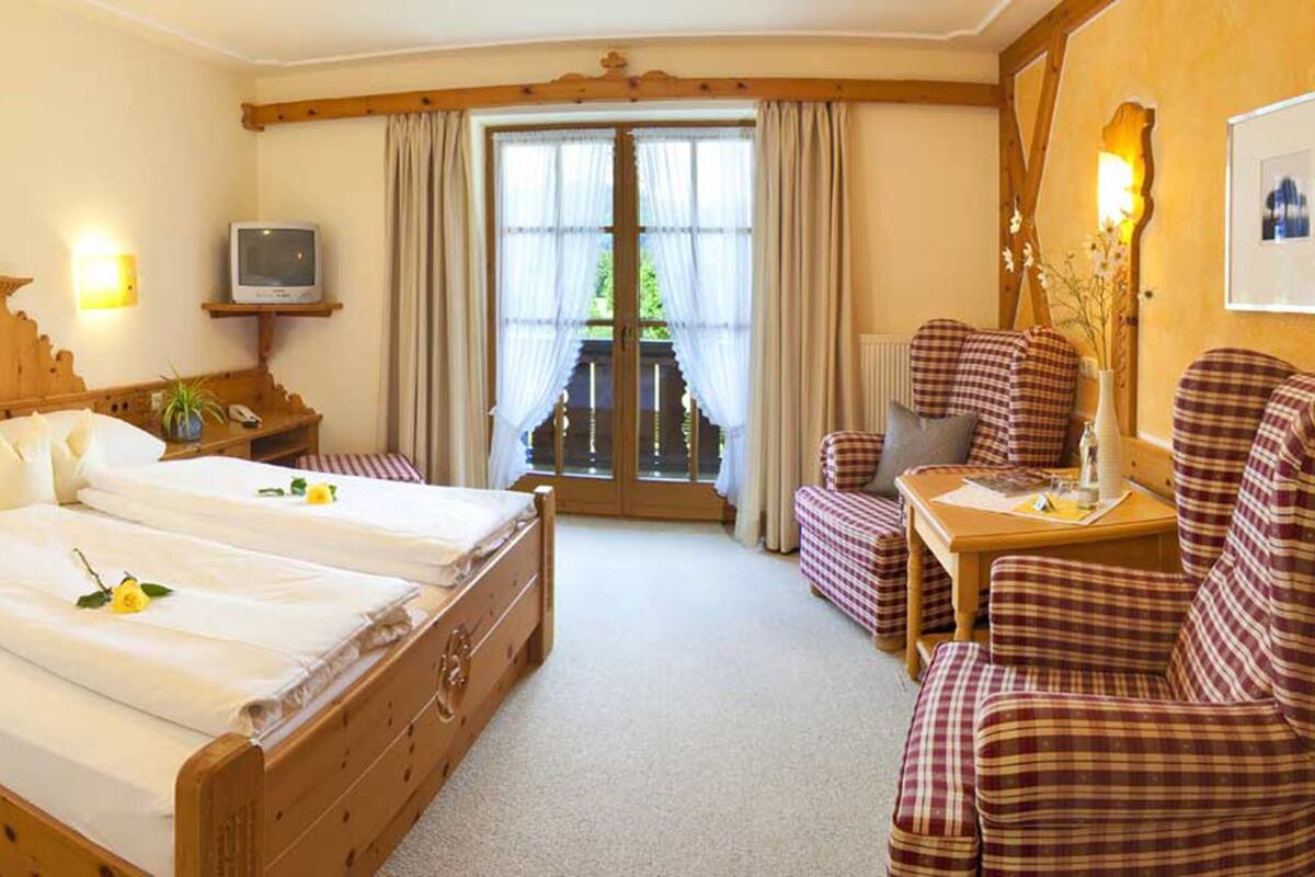 A hotel room with a wooden bed, a television, a balcony with curtains, and checkered armchairs.