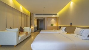 Habitación de hotel con una cama grande, un sofá y una pequeña mesa. Hay una planta en la pared y una puerta que da al pasillo.