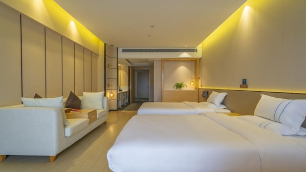 A hotel room with a large bed, a sofa, and a small table. There is a plant on the wall and a door leading to the hallway.
