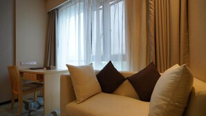 Habitación de hotel con sofá color arena, dos almohadas, escritorio de madera y ventana con cortinas translúcidas.
