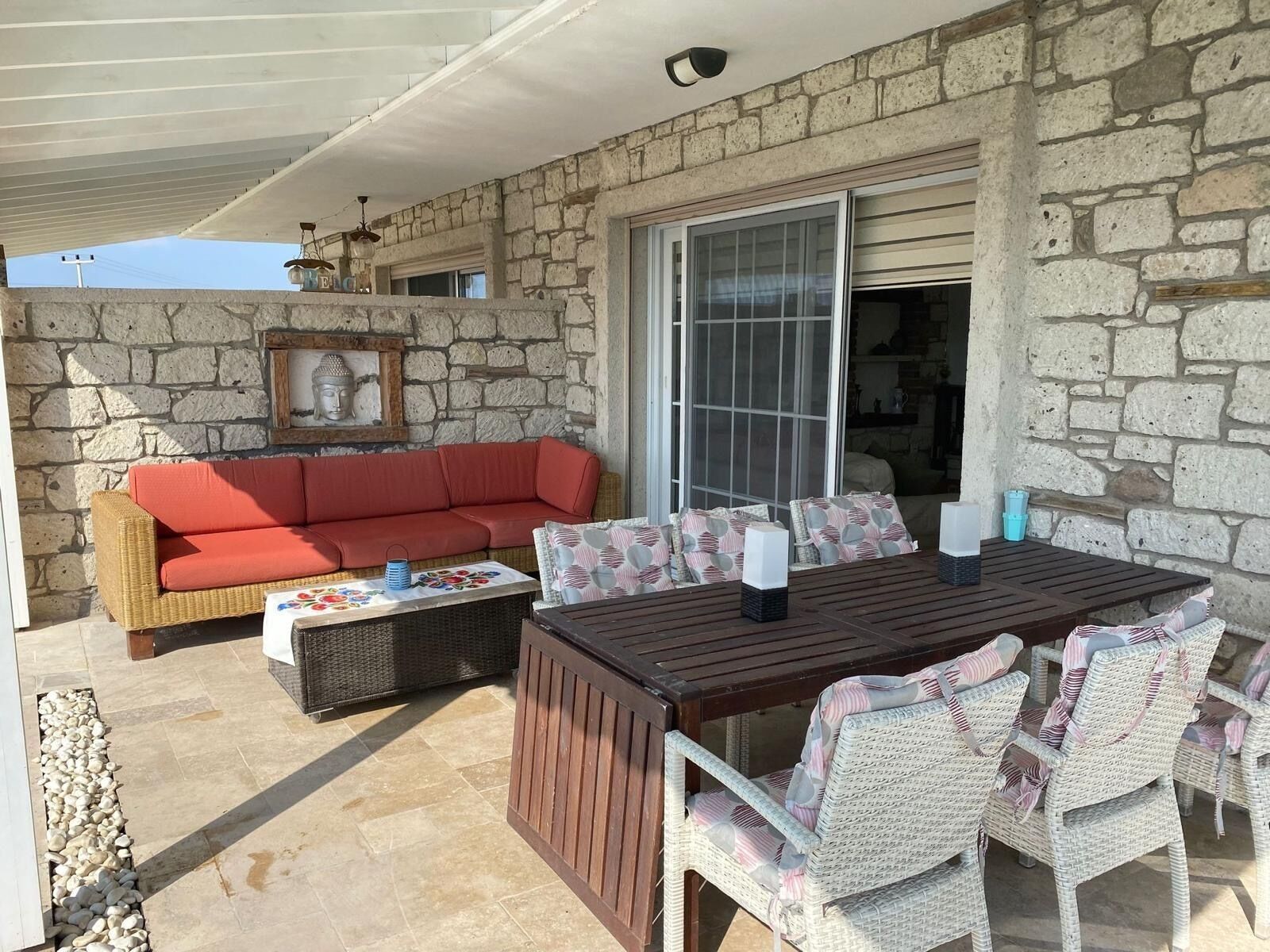 Un espacio de patio cubierto con pared de piedra, un sofá rojo, sillas de mimbre, una mesa de madera y una puerta de vidrio que da al interior.
