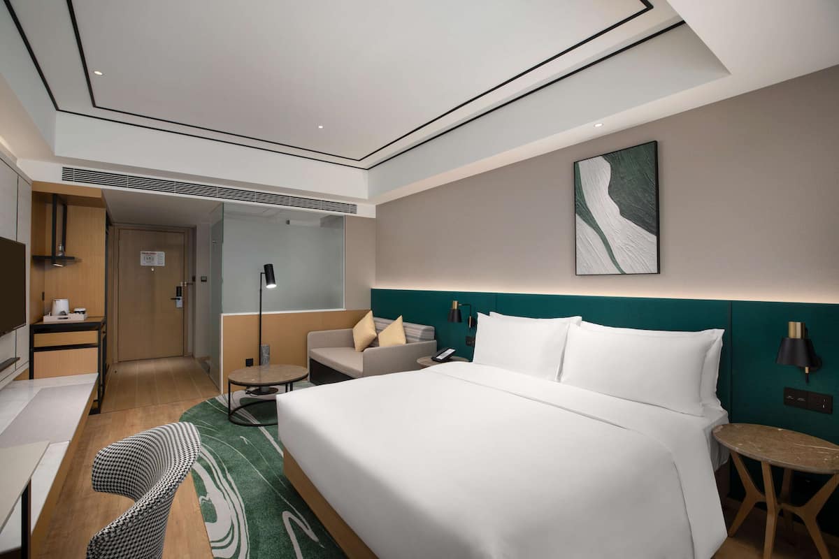 A modern hotel room with a large bed, a desk, a chair, a small table, and a wall-mounted lamp.