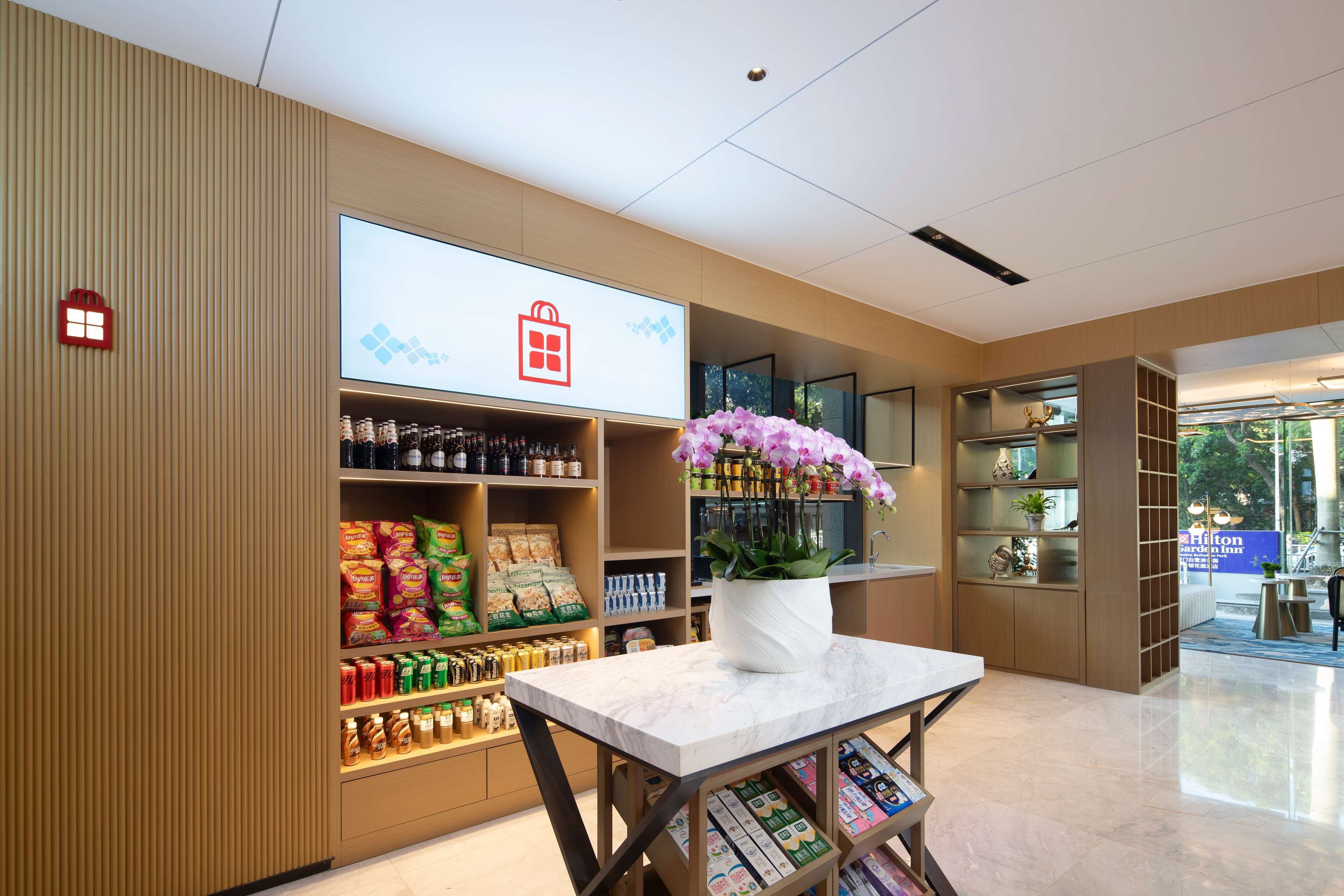 Un vestíbulo de hotel con un mostrador de recepción, una pantalla grande mostrando el logotipo del hotel, estantes con aperitivos y bebidas, y una planta en maceta con flores moradas.