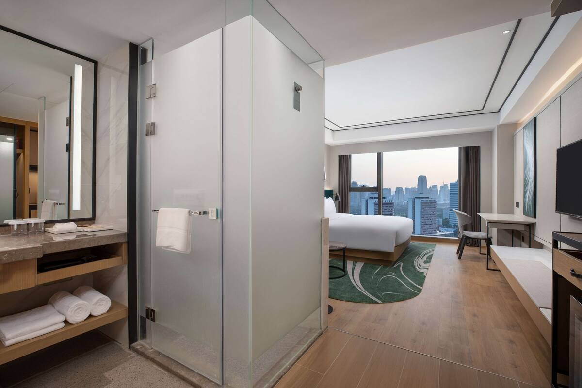 A modern hotel room with a city view, a large bed, a desk, and a shower area.