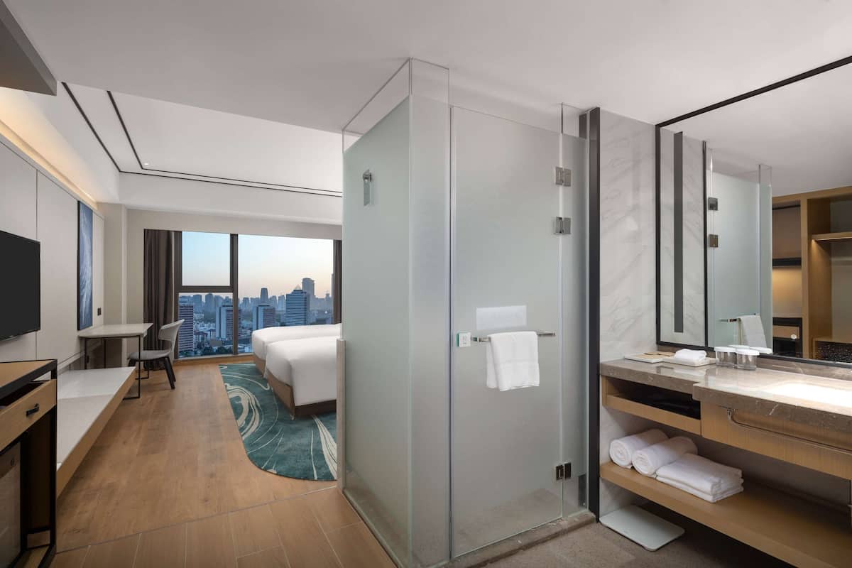 A modern hotel room with a city view, a desk, a bed, a shower area, and a bathroom with a vanity and mirror.