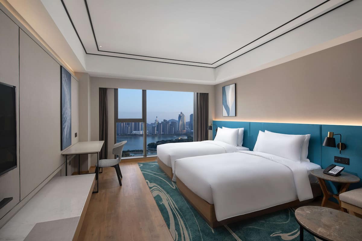 A hotel room with two beds, a desk, a chair, a small table, and a view of a cityscape.