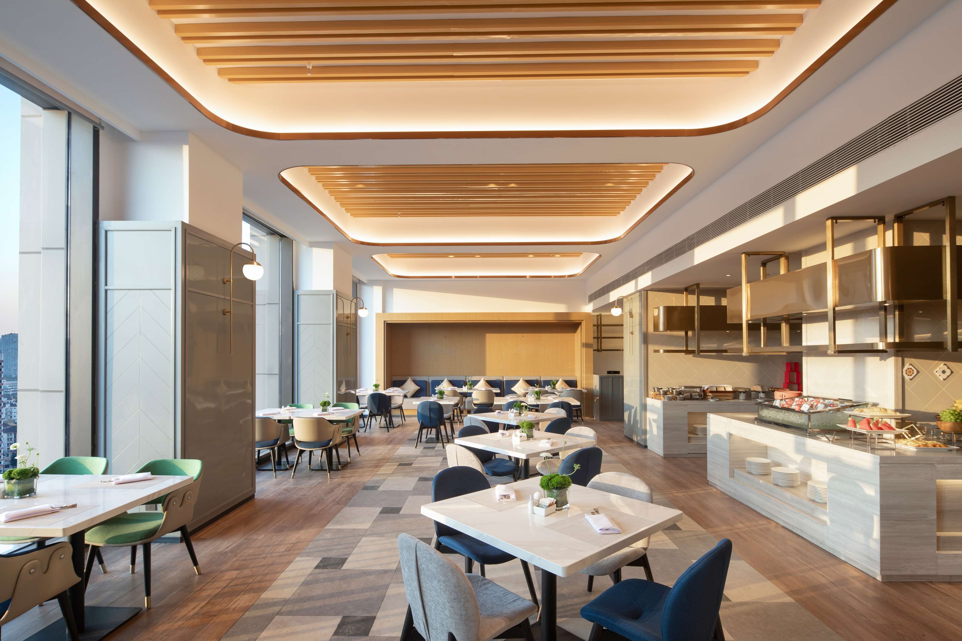 A modern dining area with multiple tables and chairs, a kitchen area with a buffet, and large windows providing natural light.