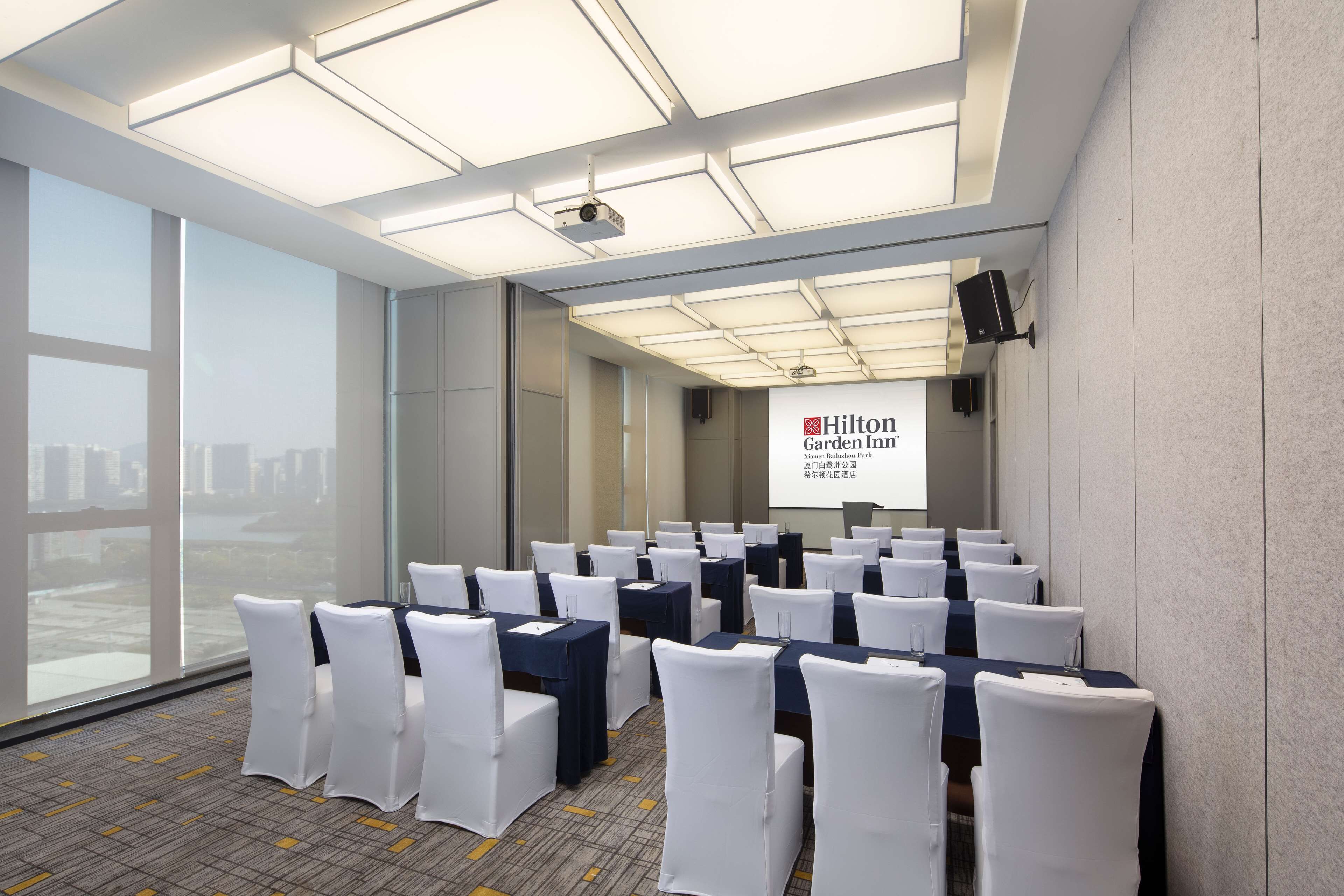 Ein Konferenzraum mit Tischen und Stühlen in Reihen, großen Fenstern und einer Leinwand, auf der das Logo des Hilton Garden Inn zu sehen ist.
