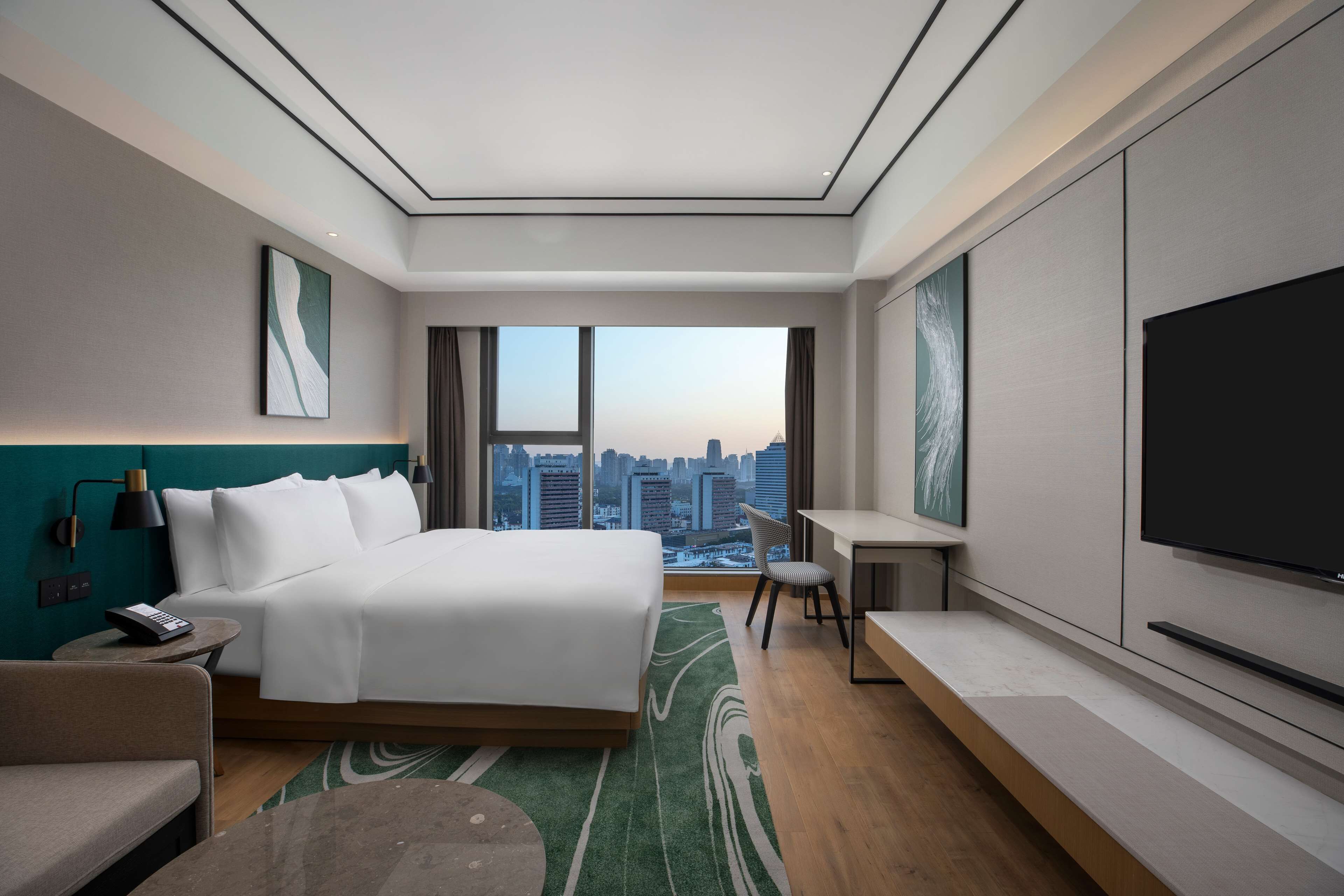 A modern hotel room with a large bed, a desk, a chair, a TV, and a view of the city skyline.