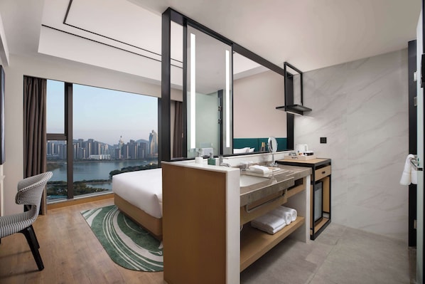 A modern hotel room with a large bed, a bathroom with a vanity, and a view of a cityscape through large windows.